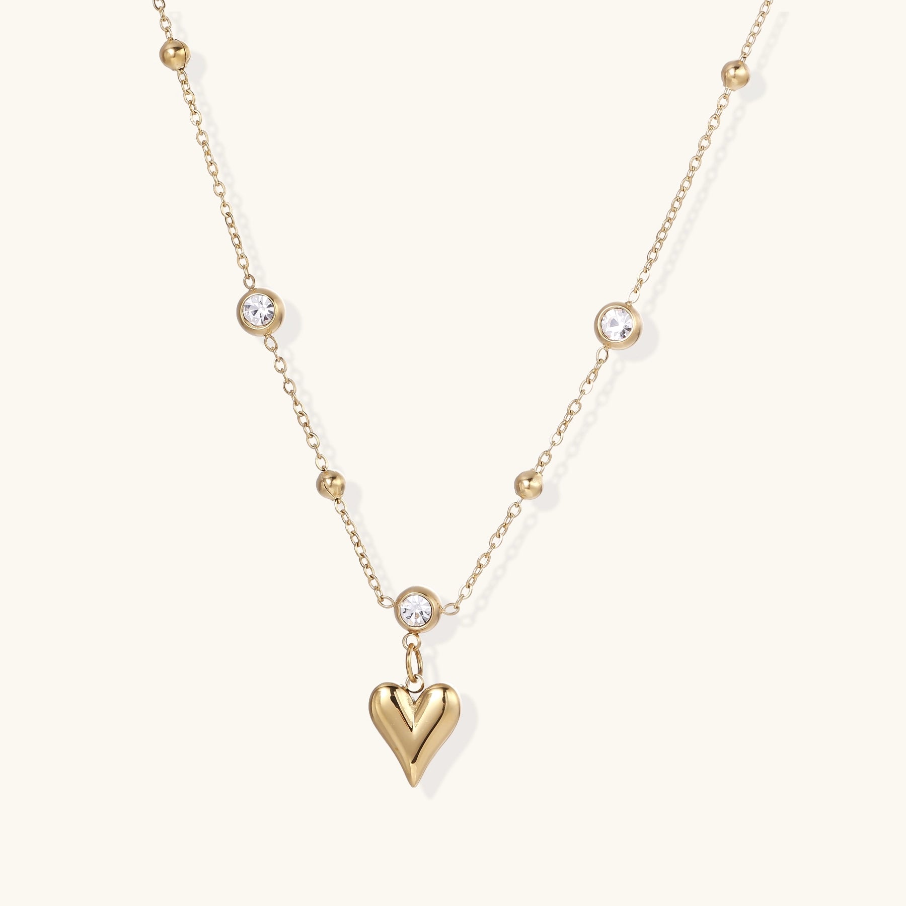 Dainty 14K gold plated necklace with heart pendant and bezel-set crystals on delicate chain against cream background.